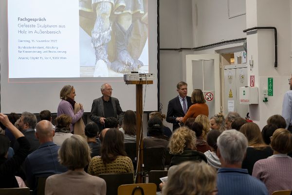 eine Gruppe von Menschen sitzt in einem Raum, vorne stehen 4 Personen und reden