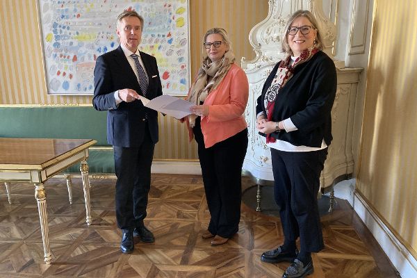 zwei Frauen und ein Mann stehen in einem Zimmer, der Mann und die Frau in der Mitte des Bildes halten gemeinsam ein Schriftstück