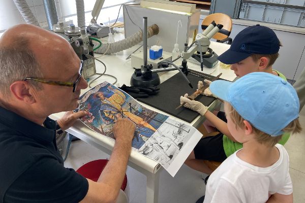 ein Mann erklärt zwei Kindern anhand eines Bildes mit einem Schweizer Söldner die Herkunft des sogenannten Schweizer Degens