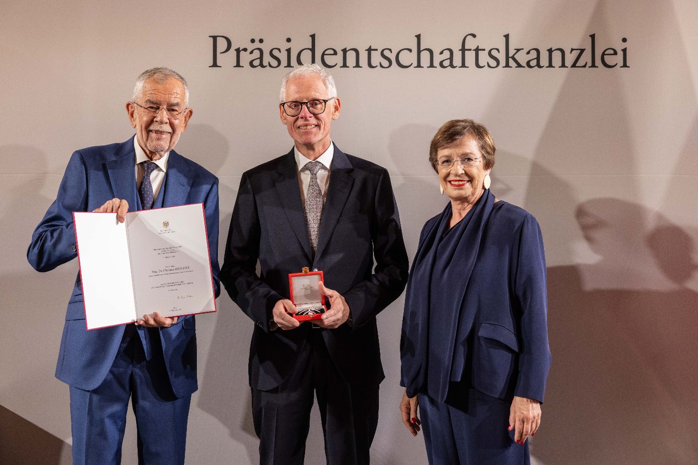Christian Brugger hält die geöffnete Ordensschatulle, links von ihm hält Bundespräsident Alexander van der Bellen die dazugehörige Urkunde aufgeklappt in die Kamera, rechts von Christian Brugger steht Doris Schmidauer, die Ehefrau des Bundespräsidenten