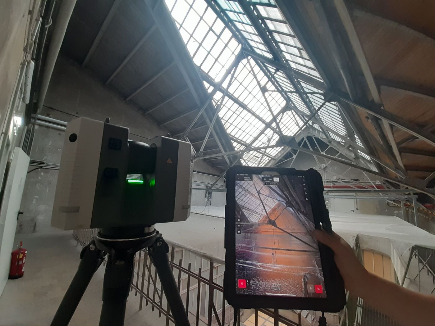 Blick in einen Dachstuhl bestehend aus einer Eisen- und Glaskonstruktion. Im Vordergrund des Bildes sind technische Geräte zur Vermessung der Dachkonstruktion zu sehen, eine Hand hält das Display eines Tablets in die Kamera