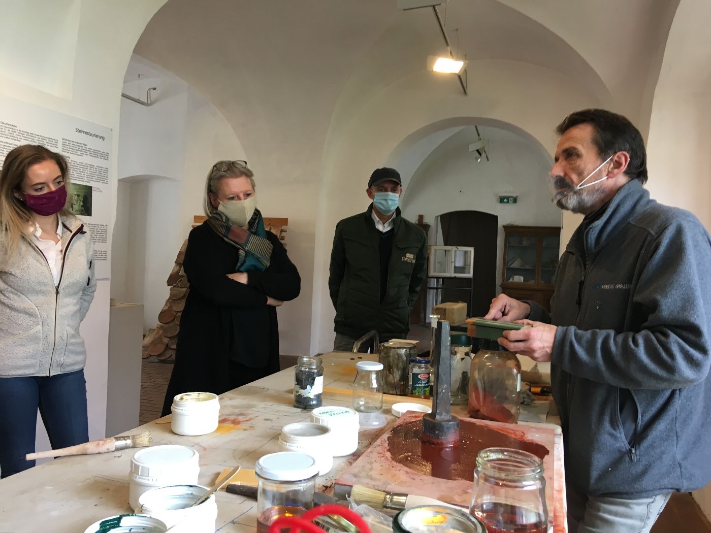 Ein Mann erklärt einer Gruppe von Besuchern in einer Werkstatt die Zusammensetzung von Farbpigmenten