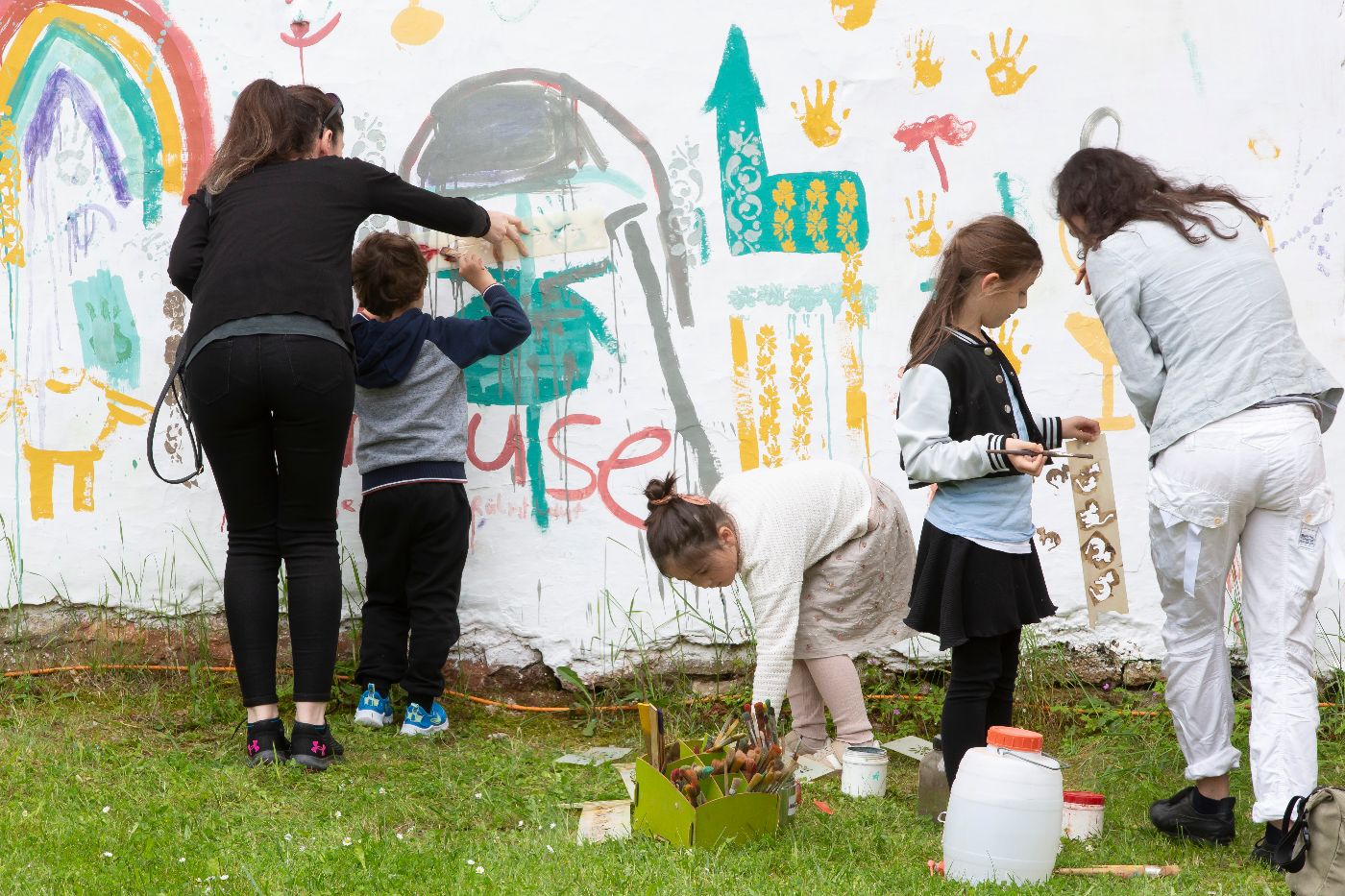 Kinder bemalen eine weiße Wand mit Farben, ein Regenbogen, färbige Handabdrücke und eine gemalte Kirche sind zu sehen