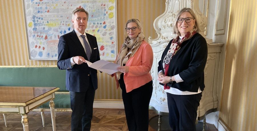 zwei Frauen und ein Mann stehen in einem Zimmer, der Mann und die Frau in der Mitte des Bildes halten gemeinsam ein Schriftstück