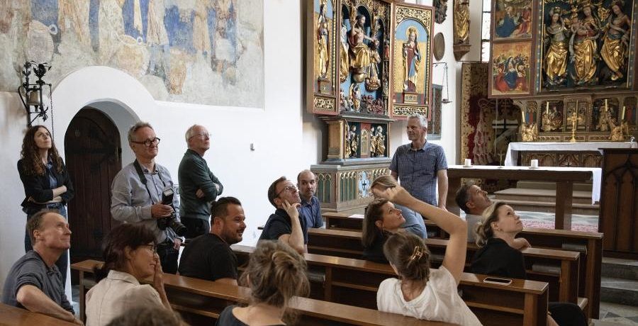 eine Gruppe von Menschen sitzt in Kirchenbänken und betrachten mittelalterliche Wandmalereien