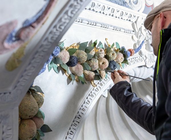 Ein Restaurator arbeitet mit Pinsel an einem Element aus Stuck, das einen Bund aus Früchten darstellt