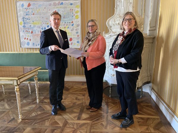 zwei Frauen und ein Mann stehen in einem Zimmer, der Mann und die Frau in der Mitte des Bildes halten gemeinsam ein Schriftstück