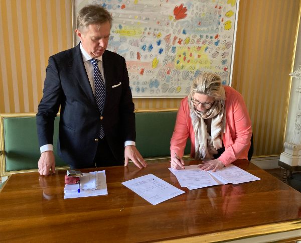 eine Frau unterschreibt an einem Schreibtisch ein Schriftstück, links neben ihr steht ein Mann und sieht zu