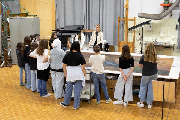 mehrere Mädchen stehen in einem Atelier um einen Tisch, auf dem die bemalte Leinwand eines Gemäldes liegt, und hören einer Frau zu die spricht