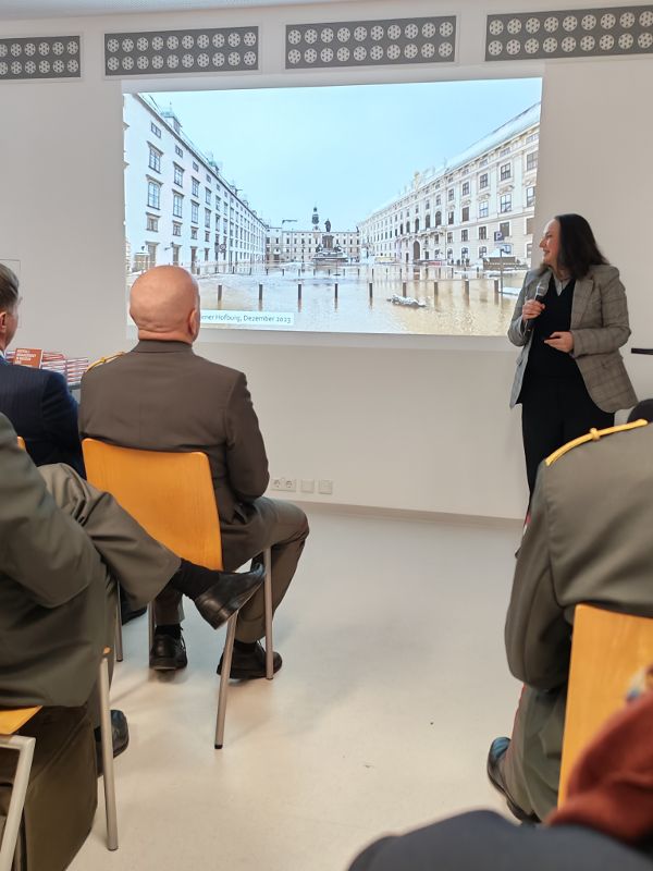 eine Frau steht in einem Vortragssaal vor einer Leinwand und spricht zum Publikum