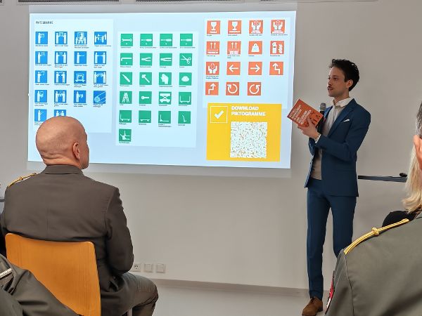 ein Mann steht in einem Vortragsaal und erläutert verschiedenfarbige Piktogramme an einer Leinwand