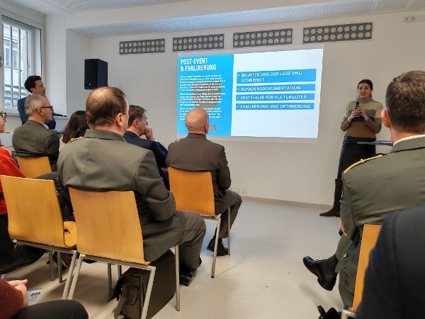 eine Frau steht in einem Vortragssaal vor einer Leinwand und spricht zum Publikum