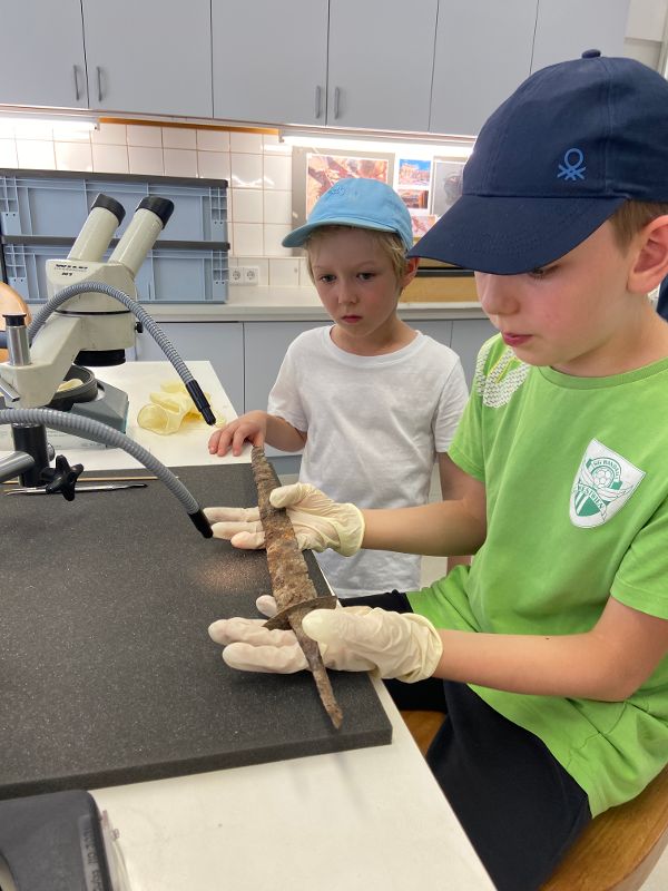 zwei Kinder sitzen an einem Mikroskop, ein Kind hält mit Latexhandschuhen bekleidet ein altes rostiges Schwert aus Eisen vorsichtig in seinen Händen