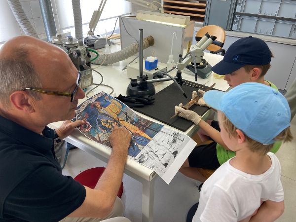 ein Mann erklärt zwei Kindern anhand eines Bildes mit einem Schweizer Söldner die Herkunft des sogenannten Schweizer Degens