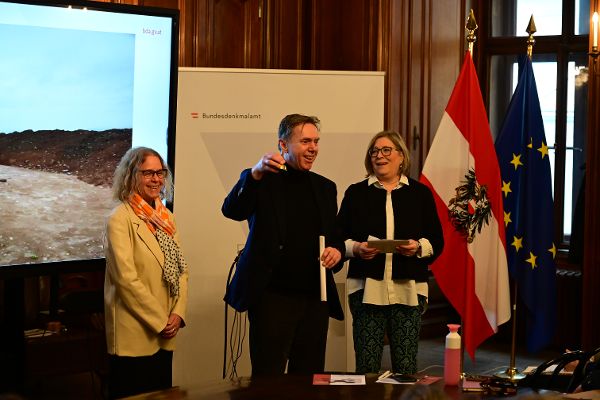 in einem Vortragssaal hält ein Mann eine kleine Glocke mit der Hand in die Höhe und spricht, zwei Frauen sehen ihm dabei zu