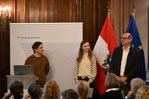 zwei Männer und eine Frau stehen in einem Vortragssaal hinter einem Rednerpult, ein Mann spricht gerade