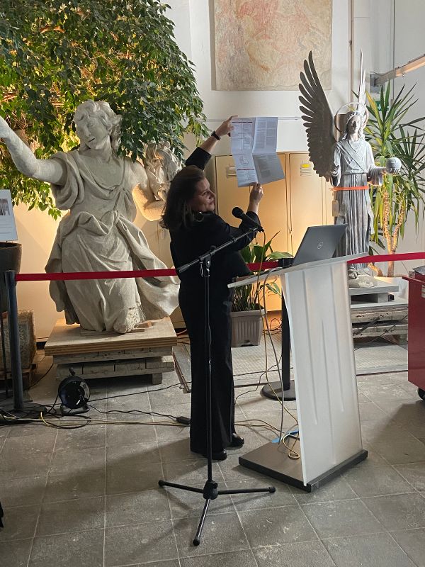 eine Frau hält ein aufgefaltetes Blatt Papier in die Höhe und spricht zum Publikum