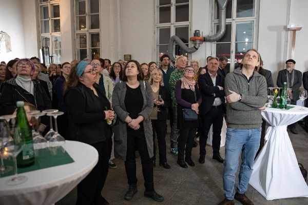 eine Gruppe von vielen Menschen steht in einem großen Saal und sieht in Richtung einer Leinwand