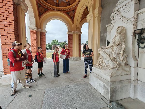 eine Gruppe von Kindern steht vor einem Grabmal mit einer Figur aus weißem Marmor, eine Frau erklärt dazu Details